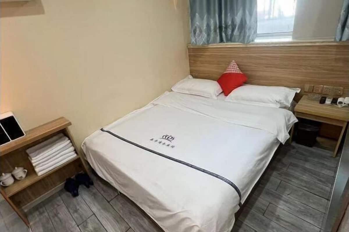 A neatly made bed with a white bedspread, a red pillow, and a wooden headboard. There is a small wooden shelf with folded towels and a pair of shoes. A nightstand with a phone and a trash bin is visible.