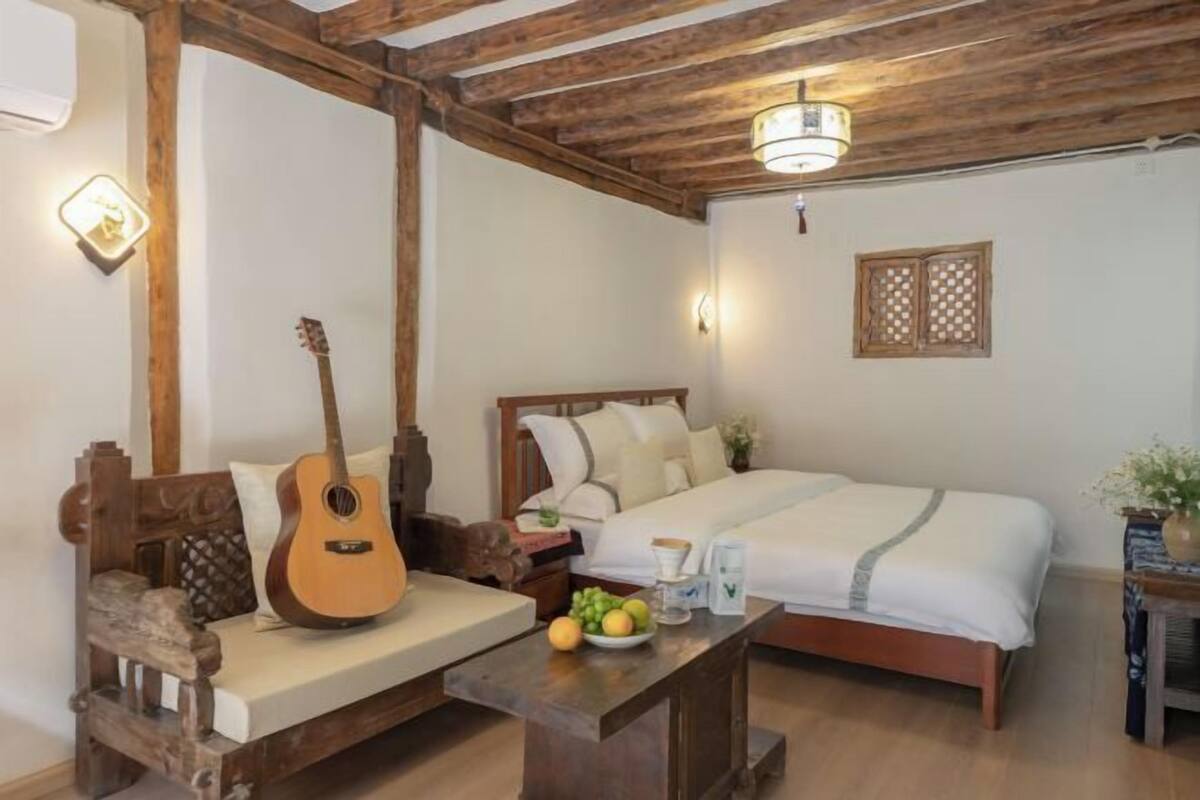 A room with a wooden bed, a wooden bench with a guitar, a small table with fruit, and a wall-mounted light fixture.