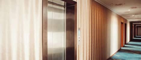 A hotel corridor with a metallic elevator, vertical wooden paneling, and patterned carpet.