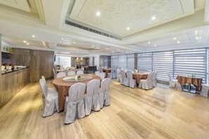 Un comedor amplio con mesas y sillas redondas, una cocina al fondo y ventanas grandes con persianas.