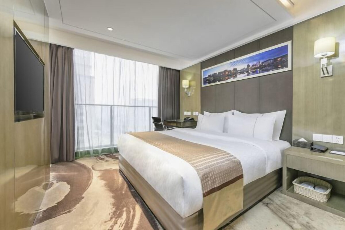 Habitación de hotel con una cama grande, un escritorio, una silla, un televisor y un cuadro con una vista urbana.