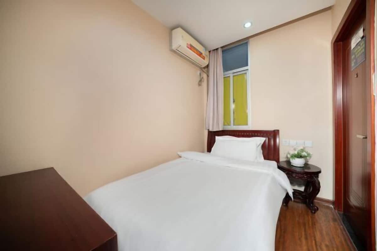 A hotel room with a bed, a wooden nightstand, a small table with a plant, an air conditioning unit, and a window with curtains.