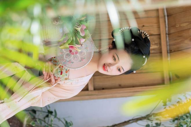 Una persona vestida con traje tradicional sosteniendo un abanico con diseños florales, en una habitación con paredes de madera y plantas a la vista.