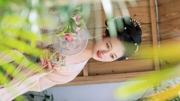 Una persona vestida con traje tradicional sosteniendo un abanico con diseños florales, en una habitación con paredes de madera y plantas a la vista.
