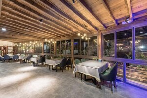 An indoor dining area with tables and chairs, wooden ceiling beams, and large windows.