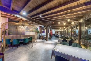 A restaurant with wooden ceiling beams, a seating area with teal chairs, and a view through large windows.