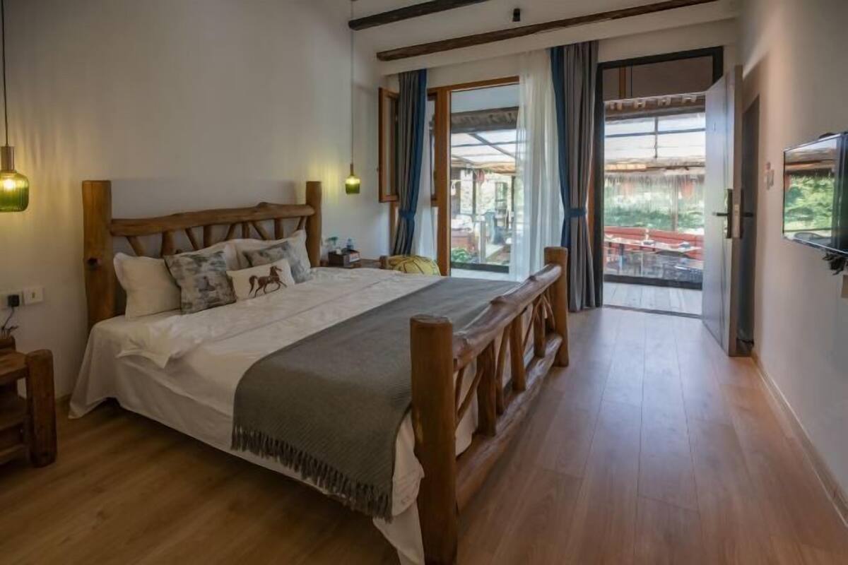 Een bed met houten frame en een witte dekbedovertrek, een houten bankje, een televisie aan de muur en een groot raam met gordijnen.