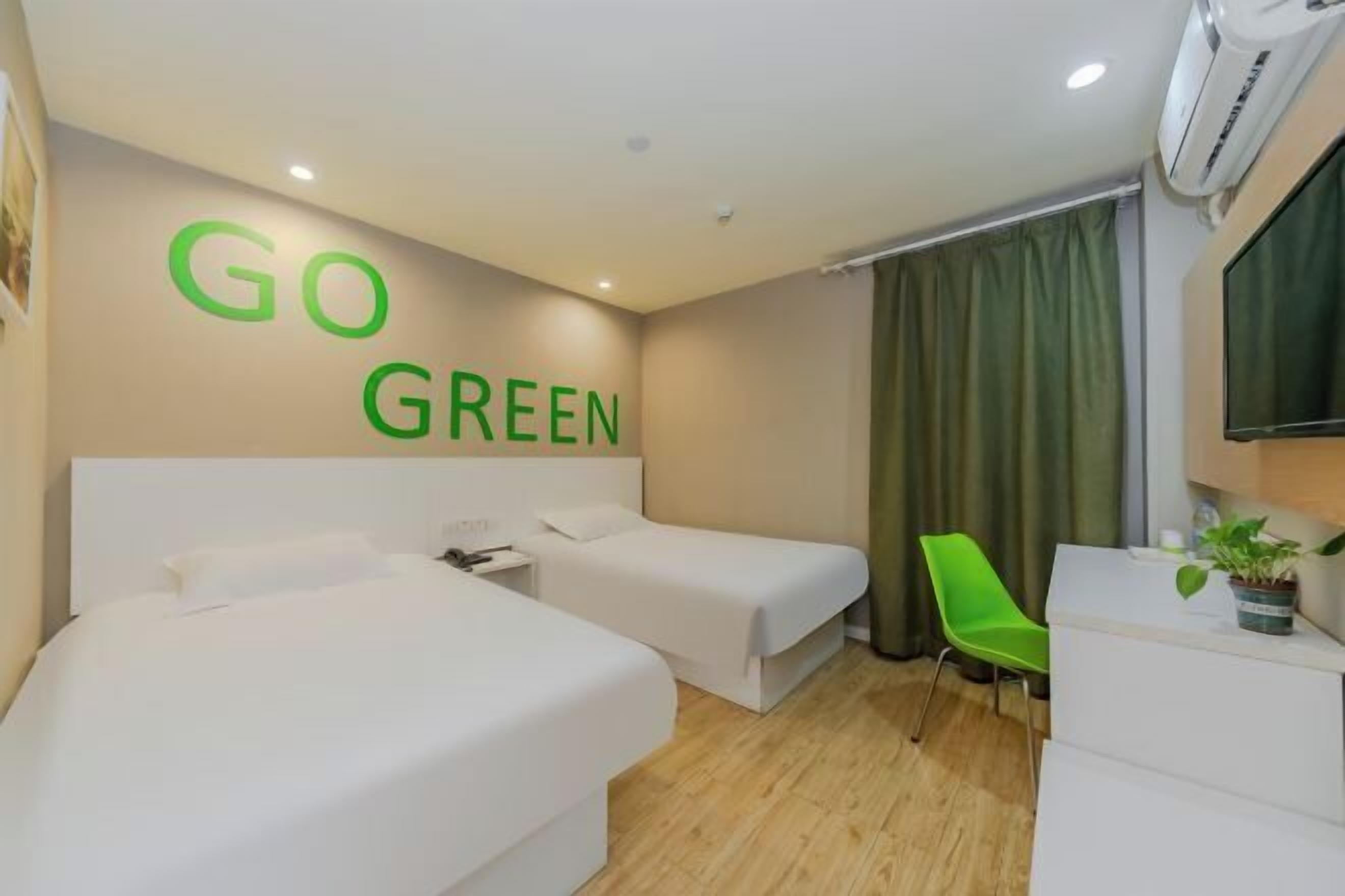 A hotel room with two beds, a green chair, a desk with a plant, and a wall with 'GO GREEN' signage.