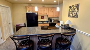 A kitchen with a granite island, wooden cabinets, a black refrigerator, a microwave, and a sink. There are three bar stools and a hanging light fixture. - Park Ave Condo #3, Sauna+Hot Tub (Park City)