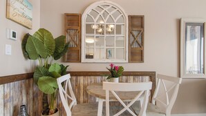 A dining area with a wooden bench, white chairs, a round table with a vase of flowers, a large window with a cross-braced design, and a wall-mounted picture frame. - Sunsets on the Gulf!  2nd Floor unit.  Available dates 8/22 - 8/31/26 (Panama City Beach)