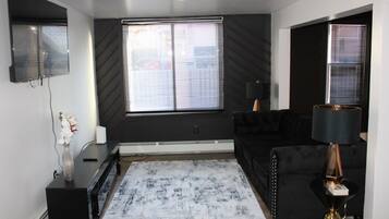 A modern living room with a large window, a black sofa, a coffee table, and a lamp.