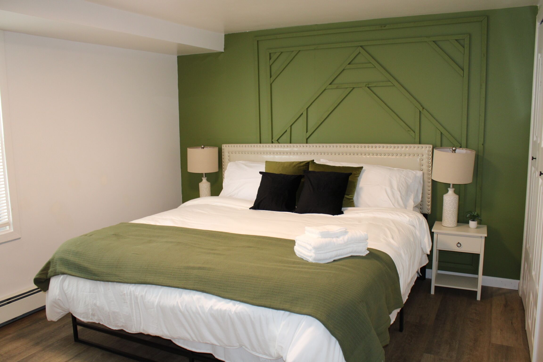 A neatly made bed with a green blanket and white linens, a bedside table with a lamp, and a green accent wall.