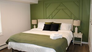 A neatly made bed with a green blanket and white linens, a bedside table with a lamp, and a green accent wall.