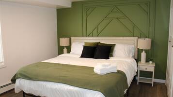 A neatly made bed with a green blanket and white linens, a bedside table with a lamp, and a green accent wall.