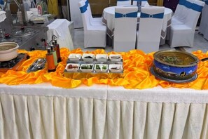 Une table de buffet garnie de divers plats et surmontée d’un grand récipient, entourée de chaises blanches ornées de rubans bleus.