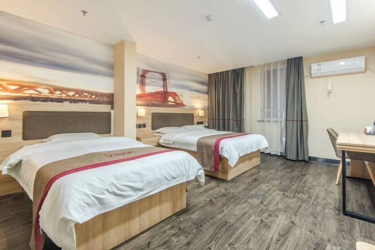 A hotel room with two beds, a wooden floor, a desk, and a window with curtains.