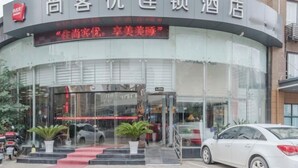 Entrada de hotel con fachada de vidrio, alfombra roja que conduce a la puerta y plantas en macetas a la entrada.