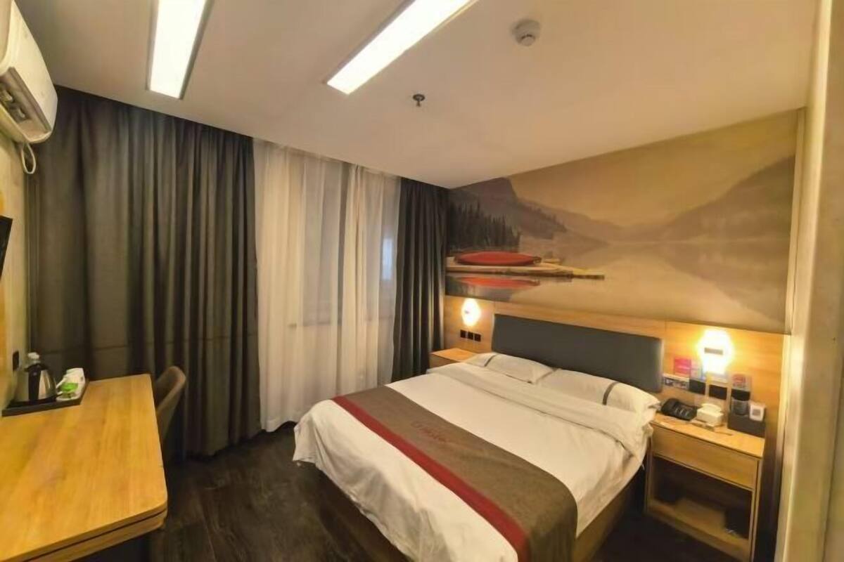 A hotel room with a bed, a desk, a chair, a nightstand with a lamp, and a painting on the wall.