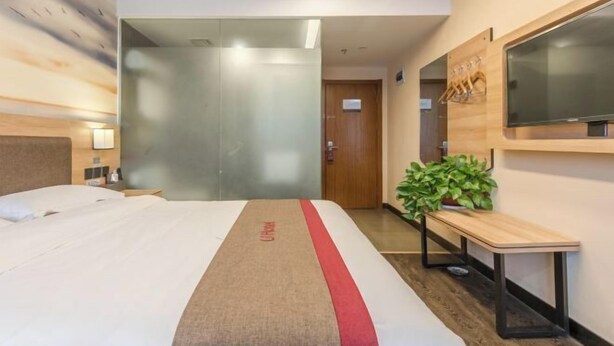 A hotel room with a bed, a TV mounted on the wall, a wooden bench, and a potted plant. - Shankee Hotel (Anqing Qijie Railway Station) (Anqing)