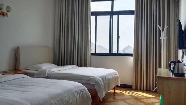 A hotel room with two beds, a window with curtains, a wooden floor, and a desk with a kettle.