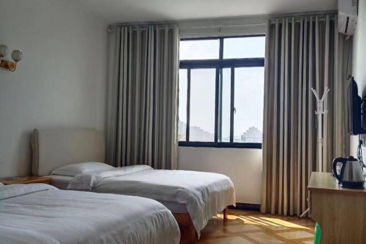 A hotel room with two beds, a window with curtains, a wooden floor, and a desk with a kettle.
