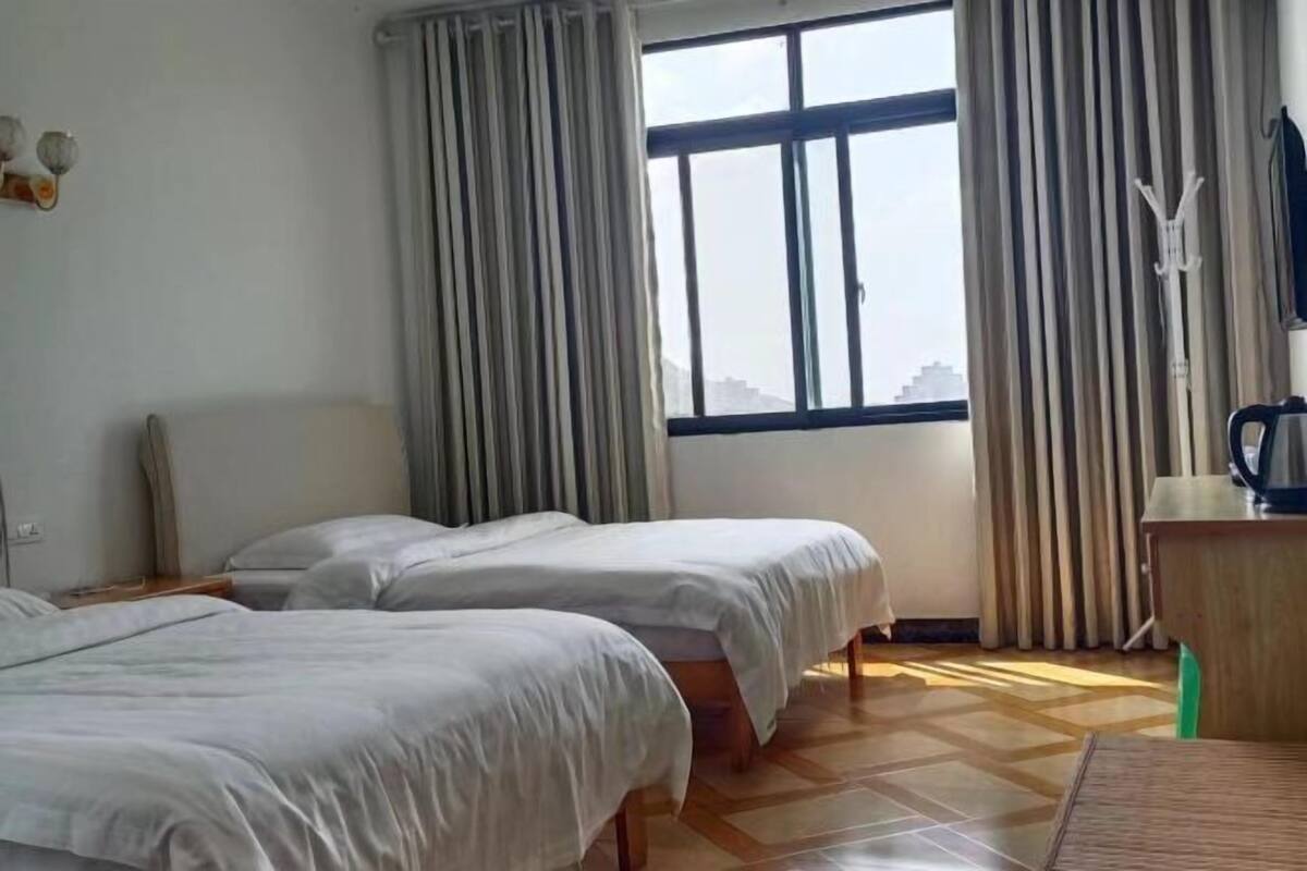 A hotel room with two beds, a window with curtains, a radiator, and a desk with a kettle.
