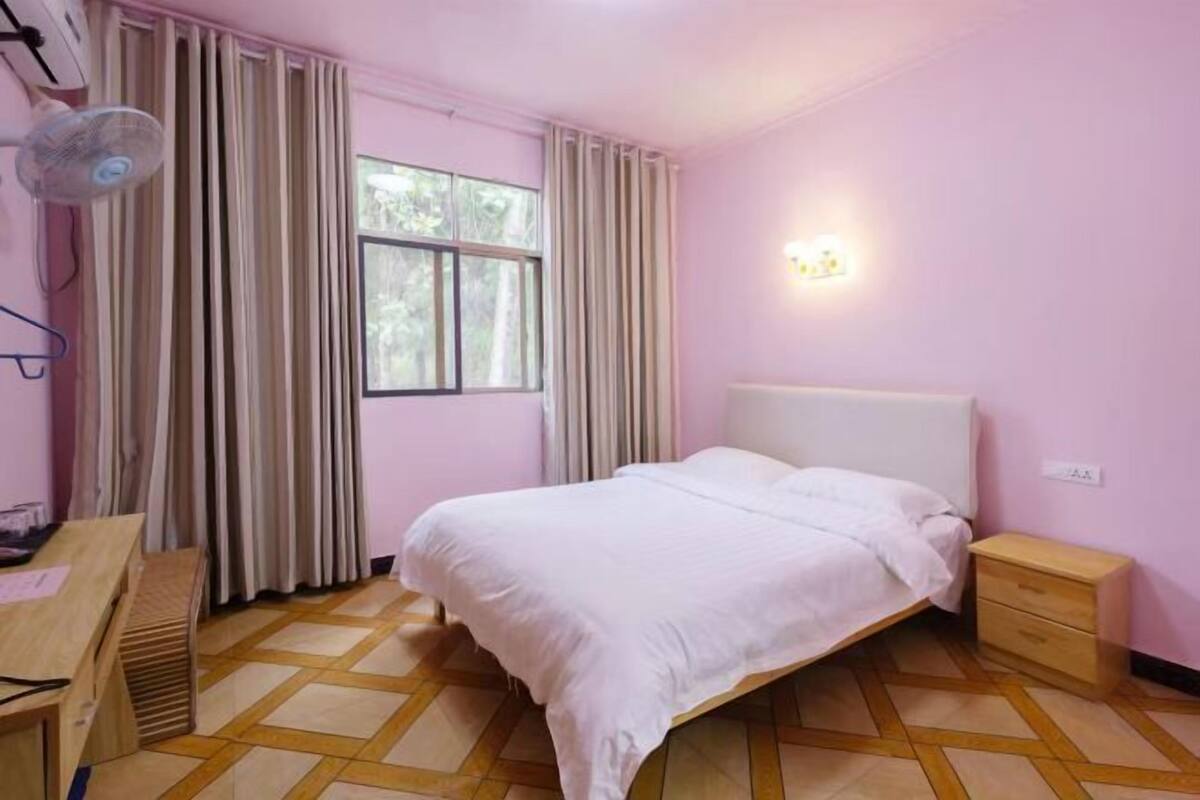 A bedroom with a bed, bedside table, a desk with a chair, a window with curtains, and a wall-mounted fan.