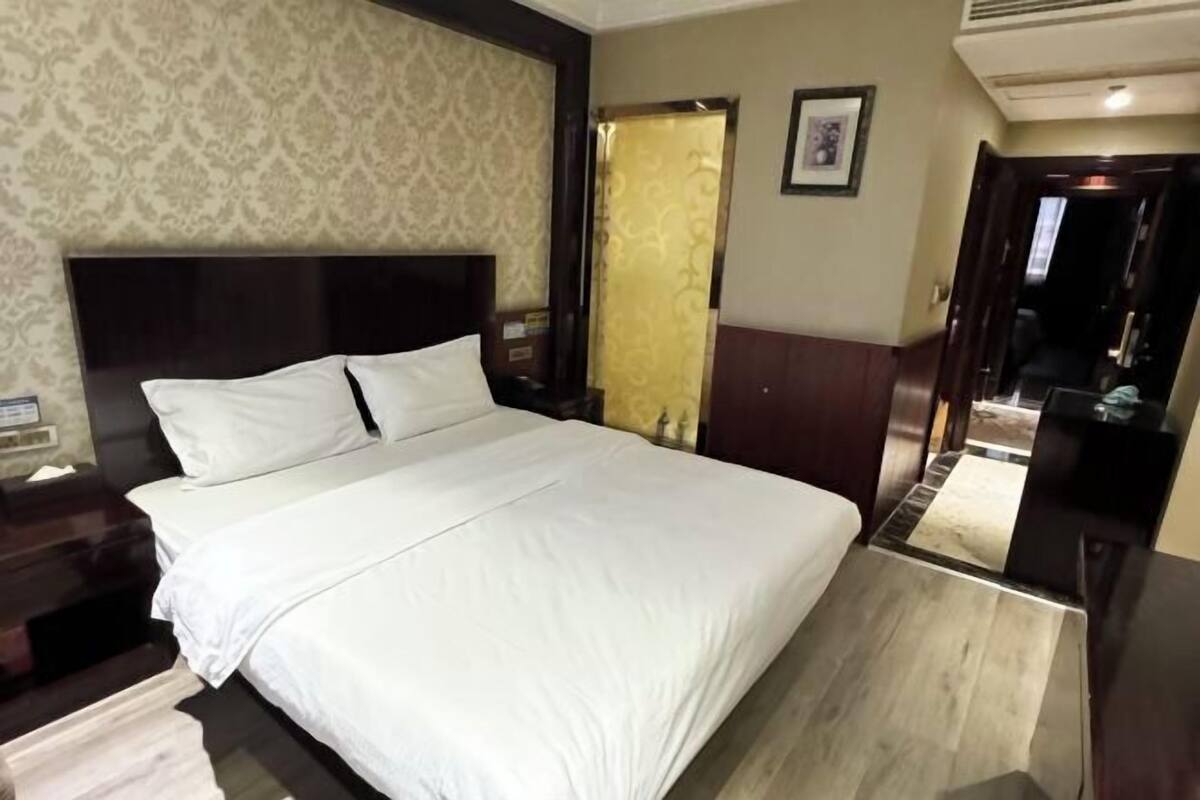 A hotel room with a large bed, a nightstand, a wooden headboard, a mirror, and a door leading to another room.