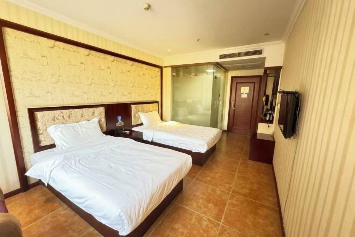 A hotel room with two beds, a television, a bathroom with a glass shower, and a door leading to another room.