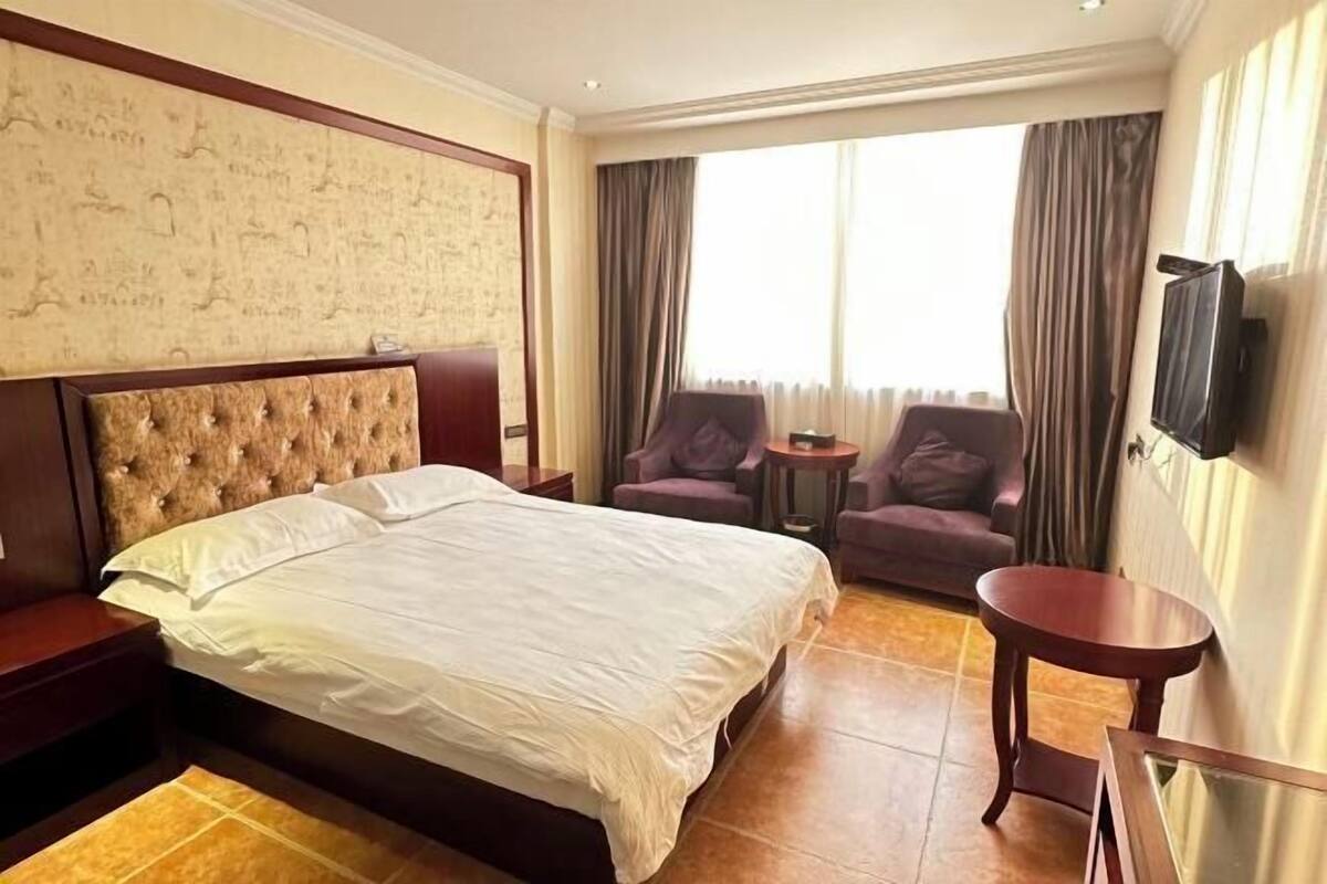 A hotel room with a large bed, a small table, two armchairs, a TV, and a large window with curtains.