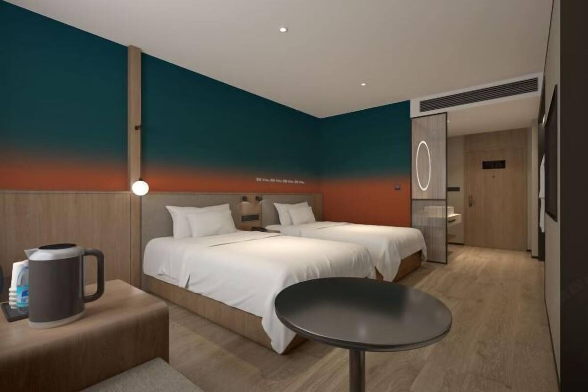 A modern hotel room with two beds, a coffee maker, a table, and a bathroom with a mirror and toilet.