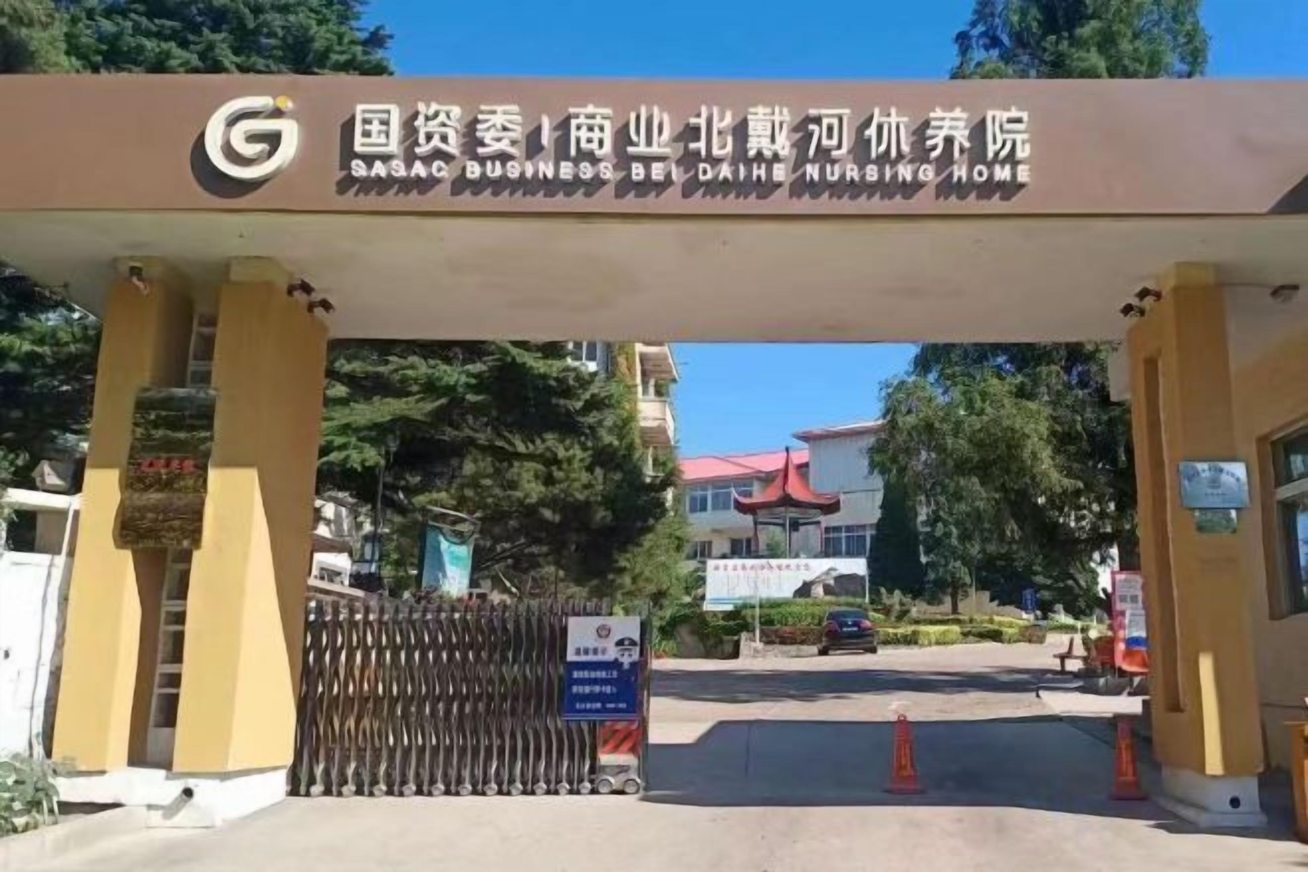 The entrance to a business hotel with a sign in a foreign language, a gated driveway, and a building in the background.