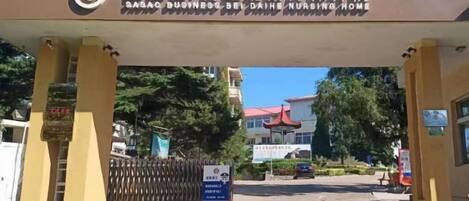The entrance to a business hotel with a sign in a foreign language, a gated driveway, and a building in the background.