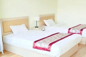 Quarto de hotel com duas camas, roupa de cama branca, mesa de cabeceira com abajur e cabeceira de madeira.