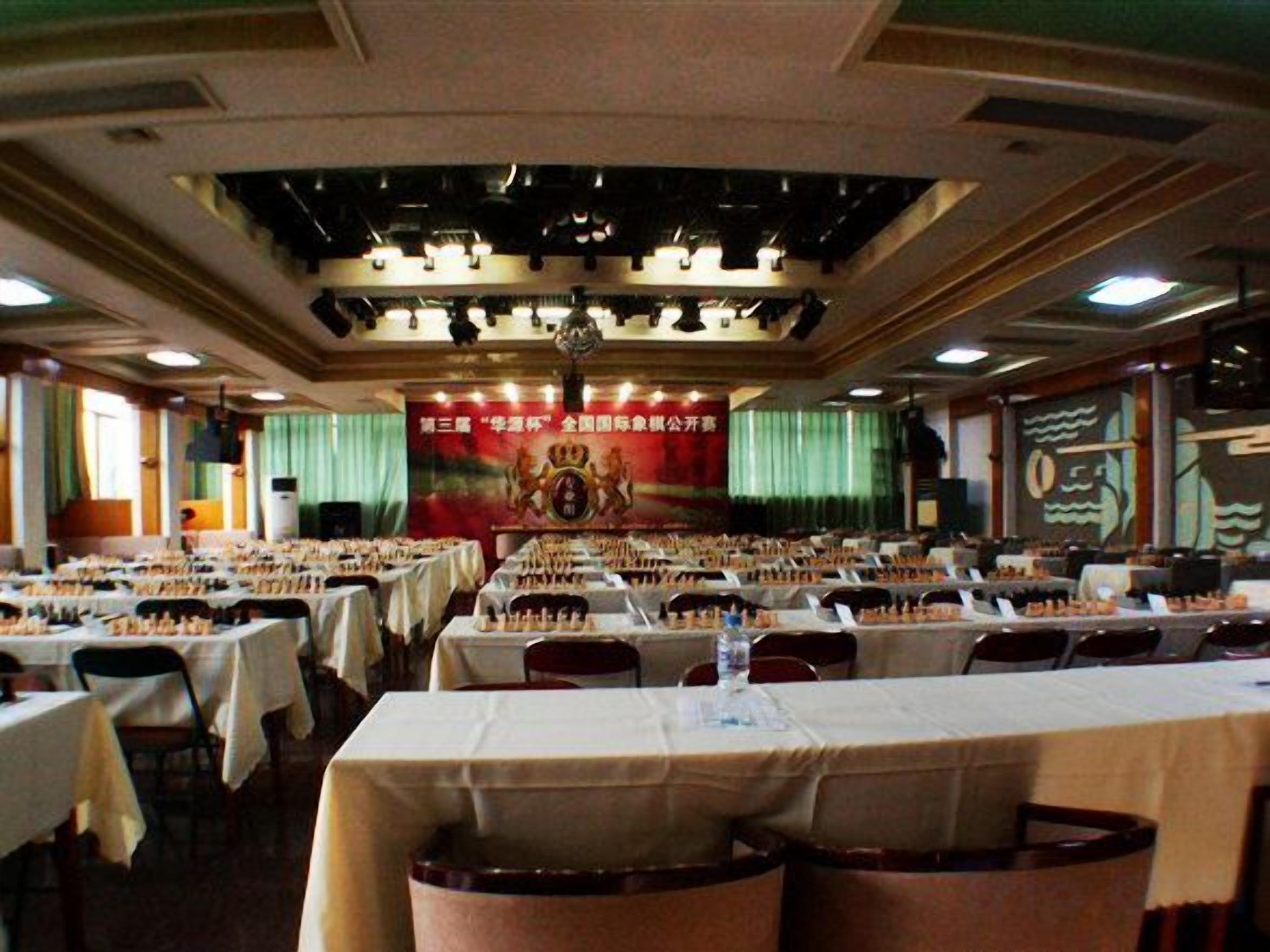 A banquet hall with multiple tables and chairs, a stage with a banner, and a chandelier.