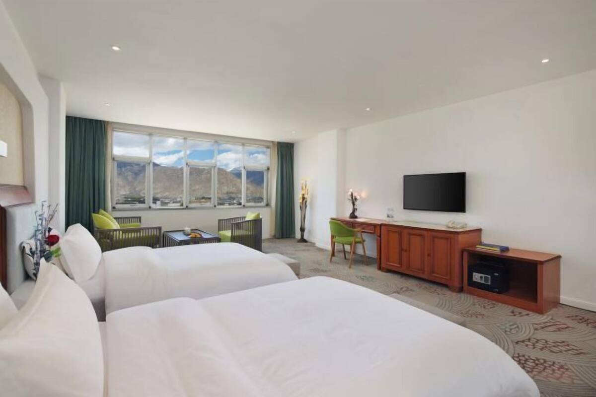 A hotel room with two beds, a desk, a television, and a large window with a mountain view.