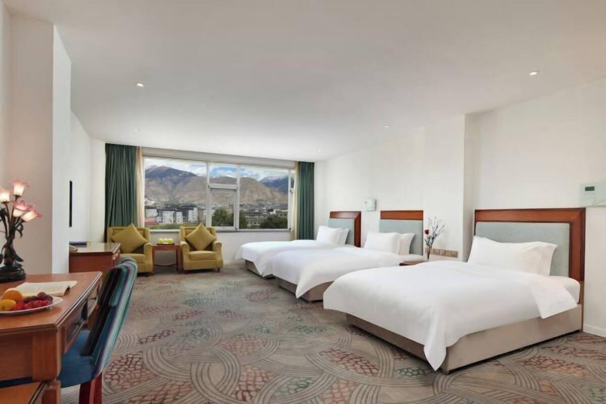 A hotel room with two beds, a desk, a chair, a window with a mountain view, and a patterned carpet.