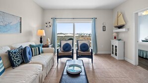 Un salón con un sofá, dos sillones, una mesa de centro, un mapa en la pared, una ventana con cortinas y vistas al mar.