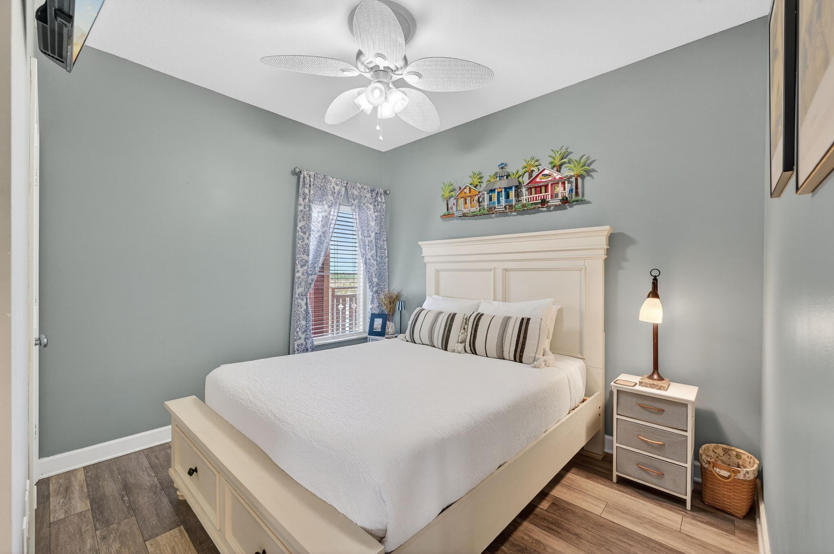A bedroom with a bed, a nightstand, a lamp, a ceiling fan, a window with curtains, and a wall decoration.