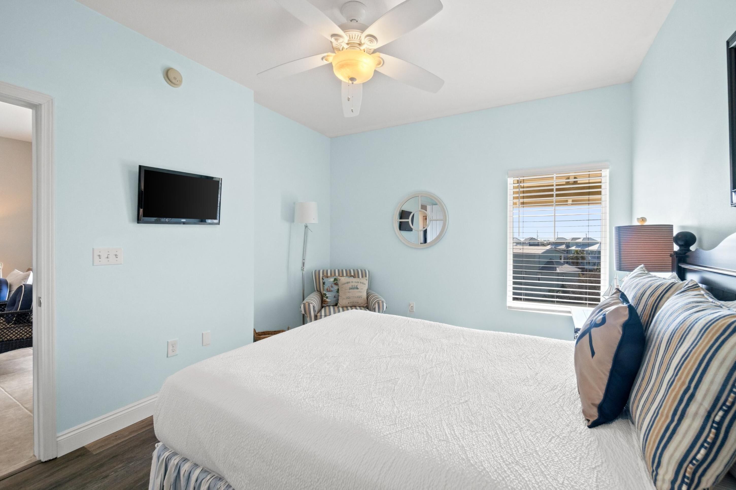 Une chambre à coucher comprenant un lit, un ventilateur de plafond, une télévision, une fenêtre avec des stores et une chaise.