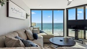 A modern living room with a large window overlooking the ocean, a ceiling fan, and a flat-screen TV.