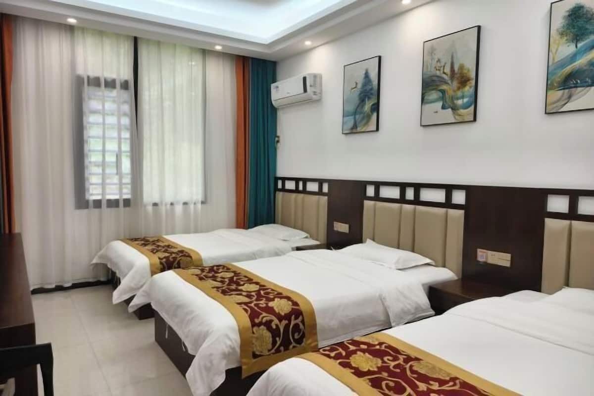 Ein Hotelzimmer mit drei Betten, einem Schreibtisch, einem Stuhl, einem Fenster mit Vorhängen, einer Klimaanlage und drei Bildern an der Wand.