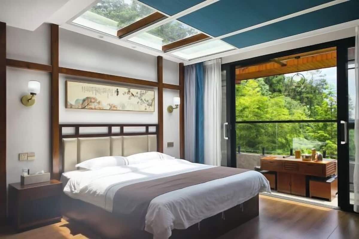 Ein Schlafzimmer mit einem Bett, einem Nachttisch, einer Wandleuchte, einem Gemälde und Blick ins Grüne durch eine Glastür.