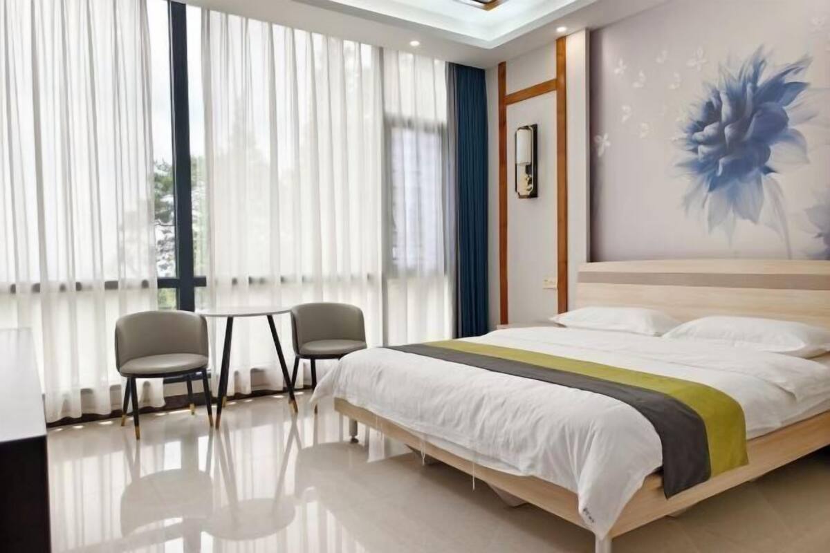 Ein modernes Hotelzimmer mit einem großen Bett, einem kleinen Tisch, zwei Stühlen und einem großen Fenster mit durchsichtigen Vorhängen.