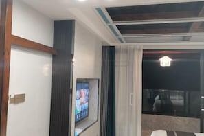 A modern hotel room with a large TV, a balcony with a view, and a ceiling with exposed beams.