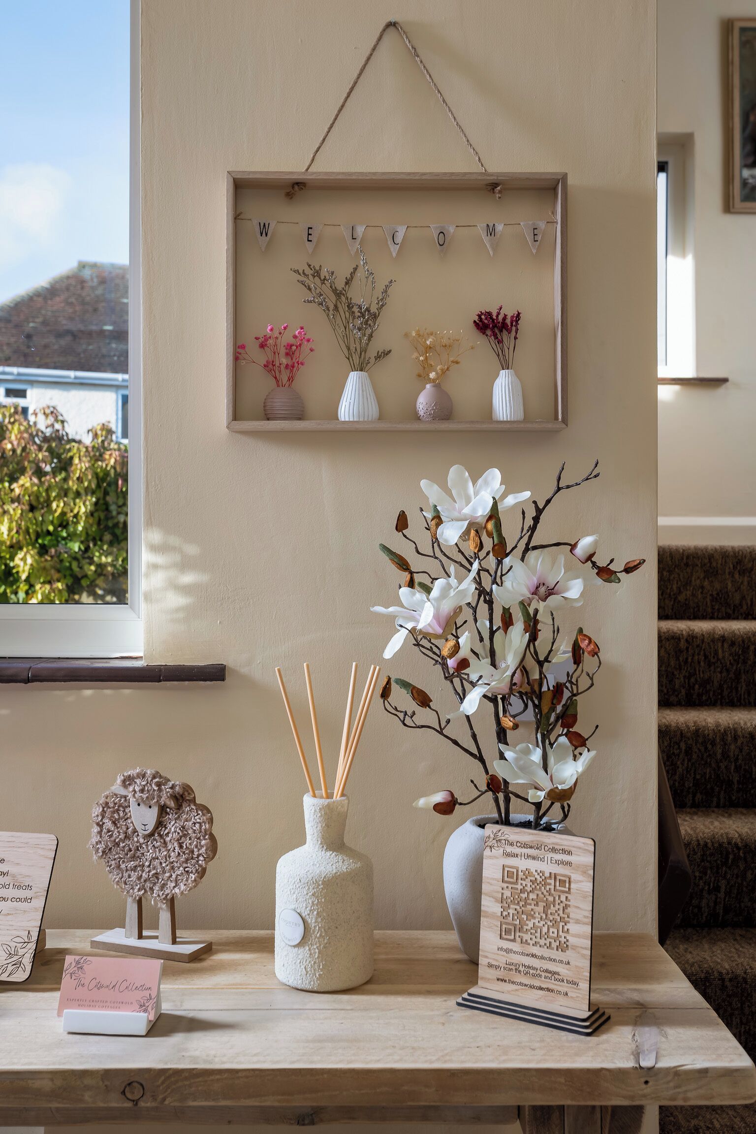 A wooden table with a framed welcome sign, a vase with white flowers, a decorative sheep, a candle holder, and a QR code sign.