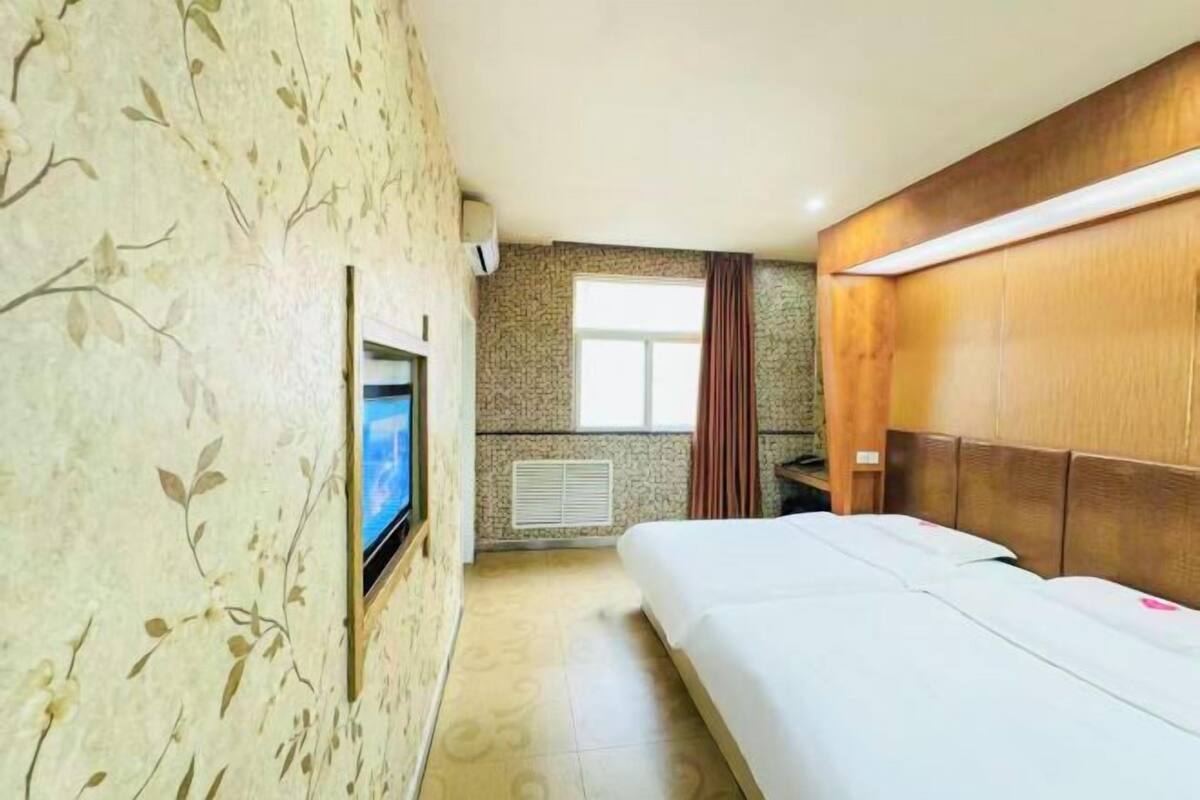 A hotel room with a bed, a TV, a window with curtains, and a wall with a floral pattern.