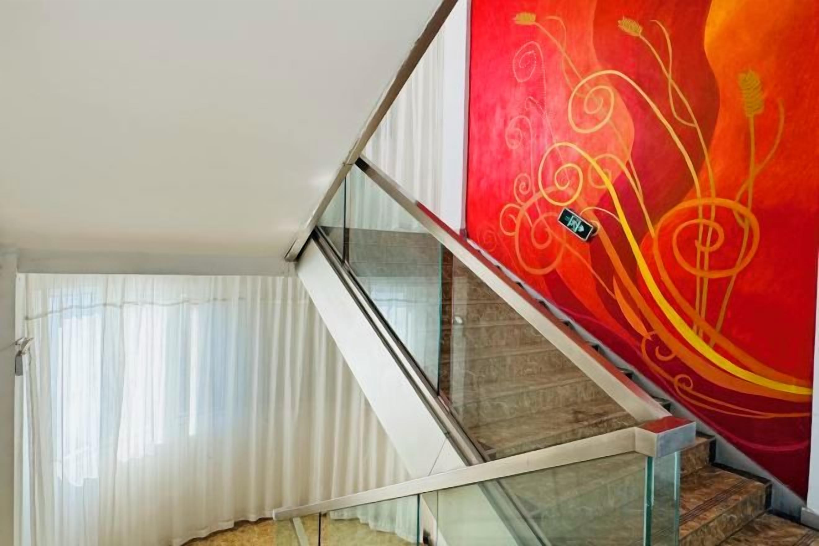 Una escalera con barandillas de vidrio, una pared roja con diseños abstractos y una cortina blanca.