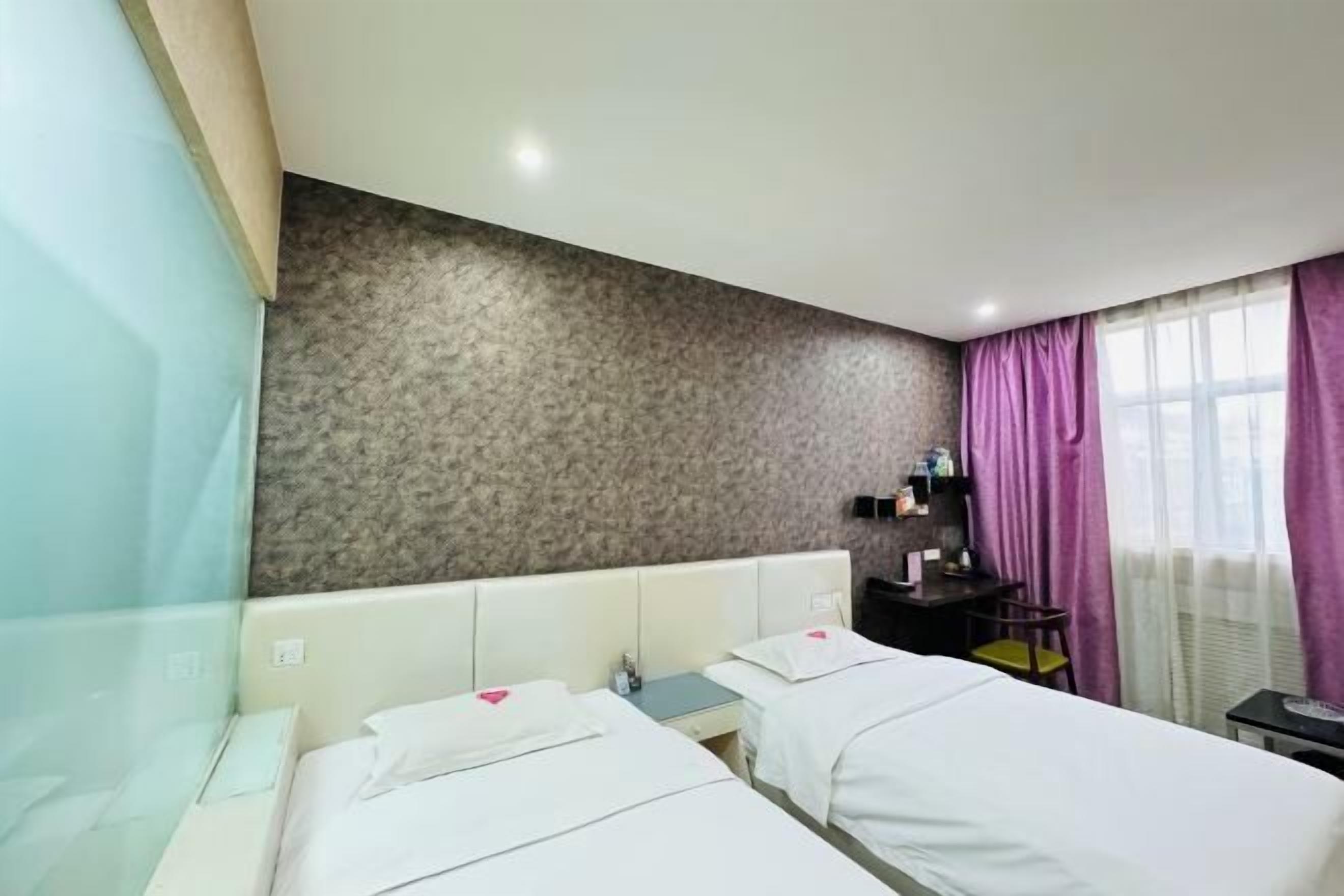Habitación de hotel con dos camas, un escritorio, una silla, una mesita y una ventana con cortinas moradas.