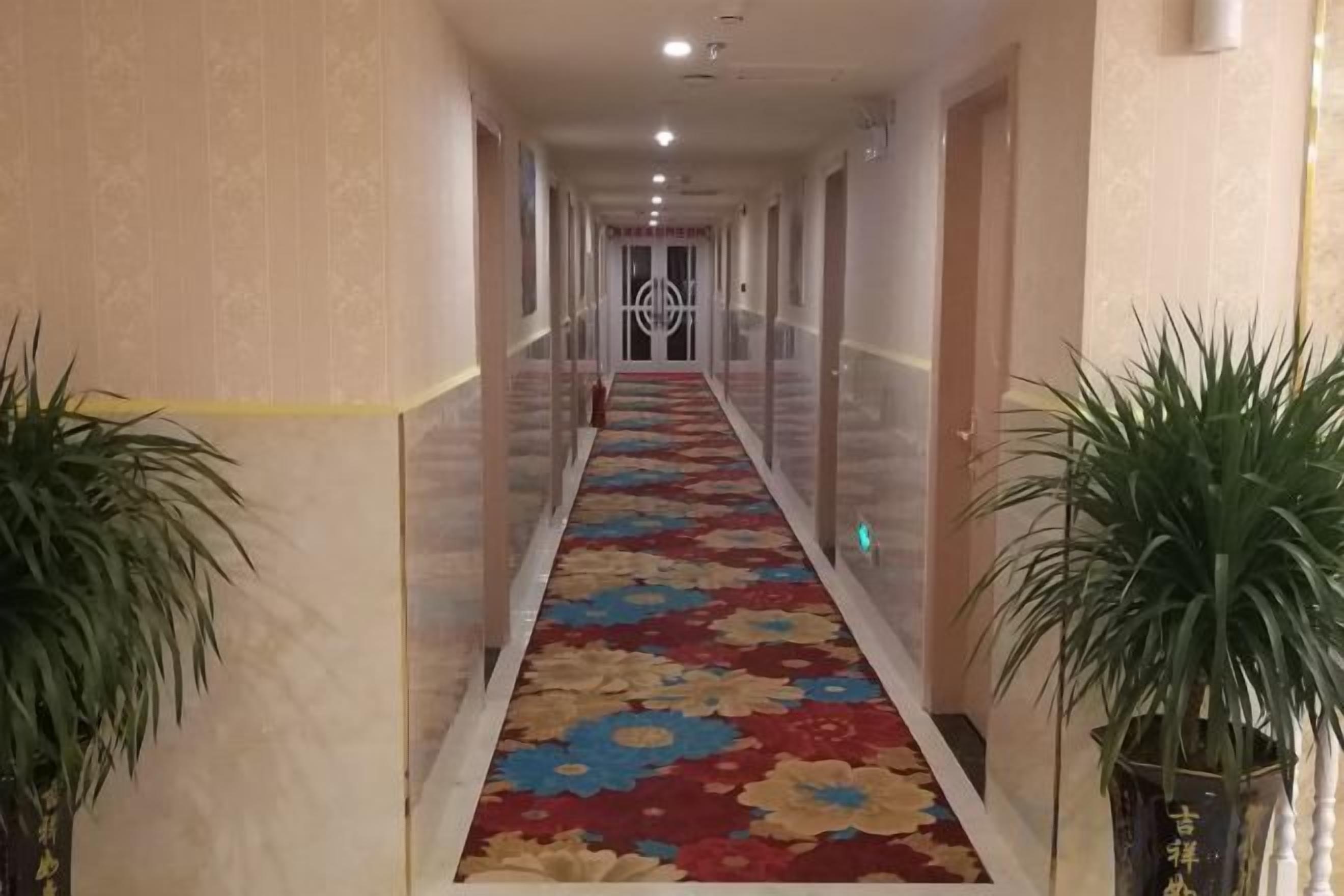 A hotel hallway with a floral carpet, potted plants, and a door at the end.